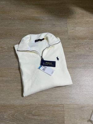 Vit halvzip från Polo Ralph Lauren - Snygg vit halvzip-tröja från Polo Ralph Lauren med klassisk broderad logga på bröstet. Tröjan är ribbstickad och har en hög krage med dragkedja. Perfekt för en stilren och avslappnad look.