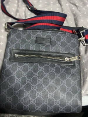 Svart axelväska från Gucci - Snygg svart axelväska från Gucci med klassiskt GG-mönster över hela väskan. Väskan har en framficka med dragkedja och en bred axelrem i rött och marinblått. Tillverkad i canvas med detaljer i läder.