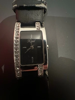 Svart klocka med stenar från Regal - Snygg svart klocka från Regal med rektangulärt ur och glittrande stenar längs ena sidan av boetten. Klockan har svart läderarmband och silverfärgad ram i rostfritt stål. Minimalistisk urtavla med diskreta markeringar och stilren design. Funkar ej!