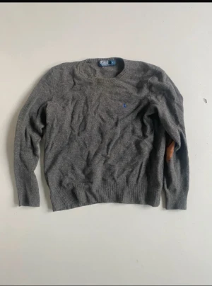 Grå stickad tröja från Polo Ralph Lauren - Mysig grå stickad tröja från Polo Ralph Lauren med rund halsringning och brunt mockadetalj på armbågarna. Liten blå logga broderad på bröstet. Perfekt för lager-på-lager och snygg till jeans.