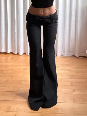 Vida svarta kostymbyxor - Säljer ett par svarta kostymbyxor med låg midja och vida ben. Byxorna har klassiska fickor och bälteshällor, perfekta för en trendig och avslappnad look. Materialet ser ut att vara en mjuk och följsam textil som ger en snygg siluett.