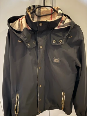 Svart Burberry regnjacka med rutigt foder - Snygg svart regnjacka från Burberry med klassiskt beige, rött och svart rutigt foder i huvan. Jackan har dragkedjor och tryckknappar framtill, två fickor med guldfärgade dragkedjor och justerbar huva. Perfekt för regniga dagar med stil.