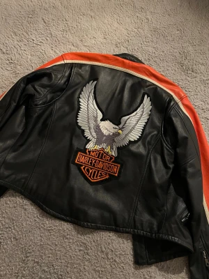 W Harley Davidson jacka (Läs beskrivning!) - Harley-Davidson skinnjacka för kvinnor (W) i Stl M. Svart med orange/vita detaljer. Klassisk bikerstil med stort broderat örnmotiv och Harley-Davidson-logga på ryggen. (som är påsytt hemma) Äkta skinn, slitstark och bekväm med snygg passform. I fint skick. Perfekt för hojåkning eller som statement-jacka i vardagen. Köpt second hand men i superfint skick (bortsett från en öppning i ena sömmen på ärmen) Se sista bilden‼️ + några enstaka fläckar på loggan där bak.