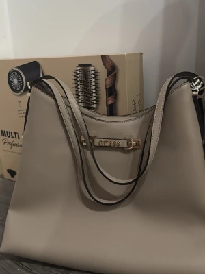 Taupe handväska från Guess - Stilren handväska från Guess i taupefärgat konstläder med guldfärgade detaljer och dragkedja framtill. Väskan är helt ny med prislapp och allt köpt för 155 euro vilket motsvarar 1700 ish 