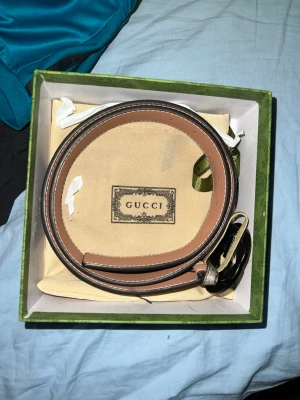 Gucci beige skärp med GG-spänne - Snyggt beige skärp från Gucci med klassiskt GG-mönster och stort silverfärgat GG-spänne. Bältet har detaljer i brunt skinn och vita sömmar längs kanterna. Perfekt accessoar för att lyfta vilken outfit som helst.