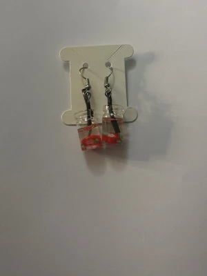 Örhängen med små burkar och jordgubbar - Unika örhängen formade som små genomskinliga burkar fyllda med miniatyr-jordgubbar i rött och grönt. Krokar i metallfärg. Perfekta för dig som gillar lekfulla och kreativa accessoarer.