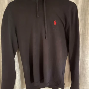 Marinblå hoodie från Polo Ralph Lauren - Marinblå hoodie från Polo Ralph Lauren med klassisk röd broderad logga på bröstet. Tröjan har huva med dragsko och är tillverkad i mjukt bomullsmaterial. Perfekt för en stilren och avslappnad look.