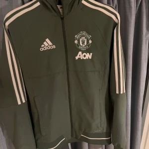 Manchester United träningsjacka adidas - Grön träningsjacka från adidas med Manchester United-logga och AON-tryck på bröstet. Jackan har vita ränder längs ärmarna, hel dragkedja och huva. Perfekt för fotbollsträning eller som sportig streetwear.