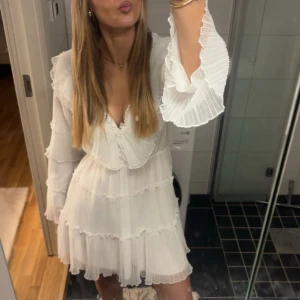 Student klänning - Superfin vit kortklänning med v-ringning och volangdetaljer. Klänningen har långa, vida ärmar med plisserade kanter och flera lager volanger för en romantisk look. Perfekt för dig som gillar en feminin och trendig stil.