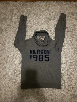 Grå Hilfiger hoodie med tryck -  grå hoodie från Tommy Hilfiger med mörkblått tryck på bröstet och snören. Storlek M, bra skick men med få fläckar som ses på bilderna. Hör gärna av dig!