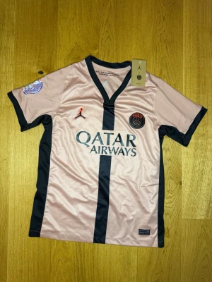 PSG matchtröja rosa Jordan Dembélé - Snygg rosa PSG matchtröja med svarta detaljer och tryck från Jordan. Framsidan har Qatar Airways-logga och PSG-emblem, baksidan har O. Dembélé och nummer 10. Korta ärmar, V-ringning och lätt, ventilerande material. Perfekt för fotboll eller streetwear.