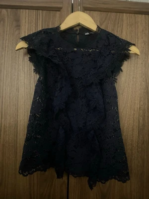 Svart spetsblus från Zara - Säljer en svart ärmlös blus från Zara i vacker blommig spets. Toppen har volangdetaljer framtill och runt axlarna, samt rund halsringning. Materialet är genombrutet och ger en elegant look. Perfekt att styla med jeans eller kjol.