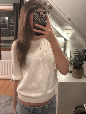 Vit stickad tröja med hålmönster - Vit stickad tröja med korta ärmar och rund hals. Tröjan har ett snyggt hålmönster upptill och ribbade muddar vid ärmslut och nederkant. Materialet känns mjukt och passar perfekt till jeans för en chill stil.