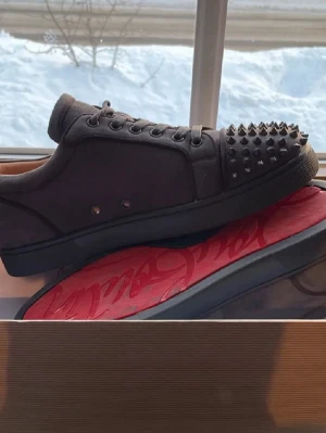 Grå sneakers med nitar Christian Louboutin - Säljer ett par gråa sneakers från Christian Louboutin med matta nitar på tån och klassisk röd sula. Skorna är låga och har snörning samt en ovandel i mocka. Perfekta för dig som vill sticka ut med en edgy och lyxig stil.