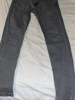 Grå Levi's raka jeans - Säljer ett par grå raka jeans från Levi's med klassisk femficksdesign och röd Levi's-tag på bakfickan. Jeansen har en avslappnad passform och är tillverkade i denim. Perfekta för en stilren och avslappnad look.