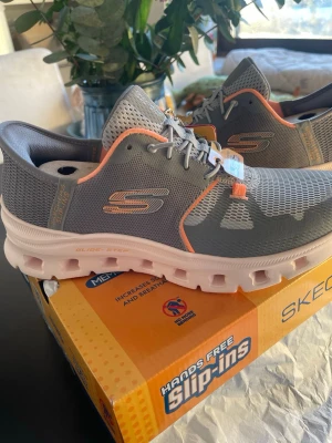 Gråa Skechers  sneakers - Snygga gråa Skechers Slip-ins sneakers med orange detaljer och vit sula. Ovandelen är i mesh för extra andningsförmåga och skon har elastiska snören för enkel påtagning. Perfekt för dig som vill ha bekväma och stilrena sneakers med modern design. Normal i storleken. Köpt för 1200kr säljes den passar inte mig stilen. Kan skickas. 