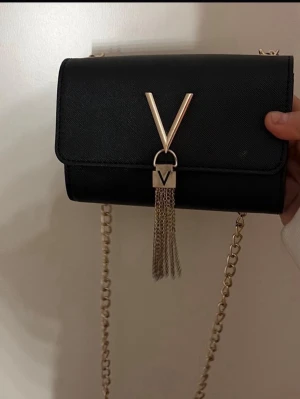 Svart axelväska med guldkedja - Snygg svart axelväska från Valentino med guldfärgad kedja och dekorativt V-spänne framtill. Väskan har en stilren, rektangulär form och är gjord i ett slätt konstläder. Framsidan pryds av hängande guldkedjor för en extra detalj.