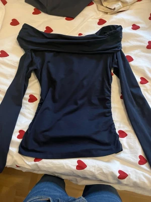 Mörkblå off shoulder topp - Snygg mörkblå långärmad topp med off shoulder-krage och figurnära passform. Toppen har rynkade detaljer vid ärmsluten och är gjord i ett mjukt, stretchigt material som känns bekvämt mot huden. Perfekt för dig som gillar en stilren och trendig look.