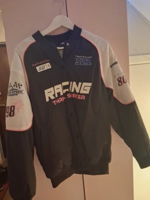 Svart racing bomberjacka - Svart bomberjacka med vita och röda detaljer samt racing-inspirerade tryck på bröst och ärmar. Jackan har ribbade muddar, knappar framtill och sportig look. Perfekt för dig som gillar streetwear och motorsport.