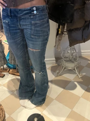 Blå bootcut jeans med fickor - Snygga blå bootcut jeans med två stora framfickor och slitningar på ena benet. Jeansen har låg midja och dubbla knappar i linningen för en cool detalj. Perfekta för en avslappnad och trendig look.