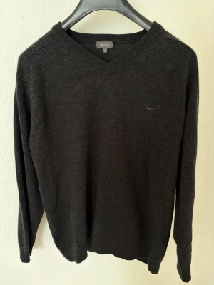 Mörkgrå stickad tröja från Wolsey - Mörkgrå stickad tröja från Wolsey med V-ringning och diskret logga på bröstet. Tröjan har långa ärmar och ribbade muddar vid hals, ärmslut och nederkant. Perfekt för lager på lager och enkel att matcha med olika stilar.