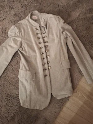 Grå kavaj från Chloe Jordache - Snygg grå kavaj med militärinspirerade detaljer och dubbla rader av silvriga knappar. Axelvaddar ger en cool siluett och fickor med lock framtill. Mjuk bomullsmix som känns bekväm och trendig.