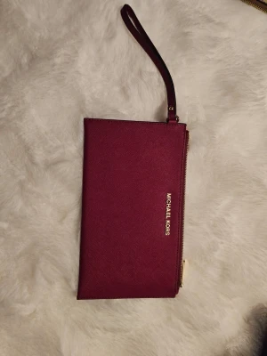 Vinröd kuvertväska Michael Kors - Stilren vinröd kuvertväska från Michael Kors i tåligt saffianoläder. Väskan har guldfärgad dragkedja, handledsrem och diskret logga framtill. Invändigt finns flera fack för kort och småsaker. Perfekt för att bära det viktigaste med stil.