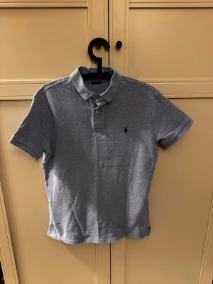 Blå piké från Polo Ralph Lauren - Klassisk ljusblå pikétröja från Polo Ralph Lauren med korta ärmar och liten broderad logga på bröstet. Slim fit-modell i mjuk bomull med krage och knappar framtill. Perfekt till jeans eller shorts.
