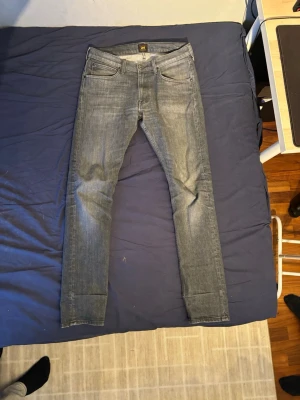 Grå slim fit jeans från Lee - Säljer ett par grå slim fit jeans från Lee med klassisk femficksmodell och snygga slitningar. Jeansen har normal midja och raka ben, tillverkade i mjuk denim med lite stretch för extra komfort. Perfekta för en avslappnad och trendig look.