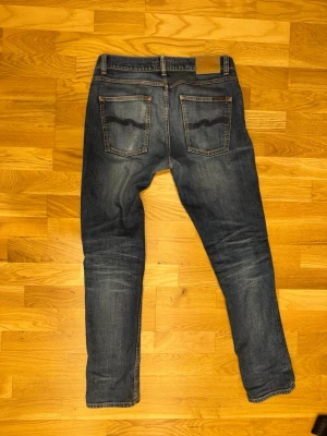 Blå jeans från Nudie Jeans 31/32 - Säljer ett par klassiska blå jeans från Nudie Jeans med snygg tvätt och kontrastsömmar. Jeansen har fem fickor, gylf med dragkedja och knapp samt Nudie-logga på bakfickorna. Materialet är slitstark denim i bomull. De har blivit lagade en gång hos nudie men det är inget som syns vid användning!
