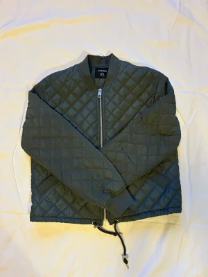 Olivgrön quiltad bomberjacka Lindex - Snygg olivgrön bomberjacka från Lindex i quiltat material. Jackan har dragkedja framtill, ribbade muddar vid hals och ärmslut samt dragsko i nederkant. Perfekt för dig som gillar en stilren och trendig look.