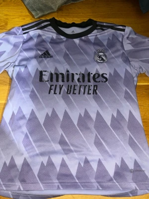 Lila Real Madrid matchtröja Adidas - Snygg lila Real Madrid matchtröja från Adidas med svart krage och mönstrad design. Tröjan har klubbmärke och Adidas-logga på bröstet samt Emirates Fly Better-tryck. Tillverkad i återvunnen polyester, perfekt för fotboll eller träning.