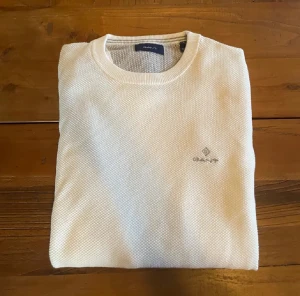 Vit stickad tröja från GANT - Vit stickad tröja från GANT med diskret logga broderad på bröstet. Tröjan har rund halsringning, ribbade muddar och långärmad passform. Materialet är mjukt och strukturerat, perfekt för dig som gillar clean och klassisk stil.