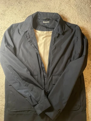 Mörkblå vindjacka från Kappahl - Snygg mörkblå vindjacka från Kappahl i storlek S. Jackan har klassisk krage, dragkedja framtill och två stora fickor. Insidan är fodrad i beige och materialet känns lätt och smidigt. Perfekt för dig som gillar stilren och enkel design.