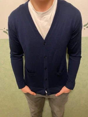 Marinblå kofta från Riley i merinoull - Snygg marinblå kofta från Riley i mjuk merinoull. V-ringad med knappar framtill och två praktiska fickor. Perfekt att slänga på över en t-shirt för en chill och stilren look. Långärmad och med normal passform.