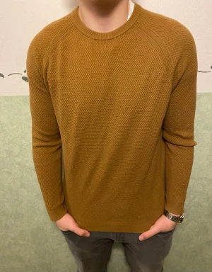 Brun stickad tröja från Riley - Snygg brun stickad tröja från Riley i storlek M. Tröjan har rund hals, långärmade raglanärmar och ett diskret texturstickat mönster. Perfekt för dig som gillar enkel och stilren design med lite extra struktur.