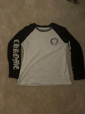 Chrome Hearts svartvit långärmad tröja - Säljer en svartvit långärmad tröja från Chrome Hearts med kontrastärmar och stora tryck på både fram- och baksida. Tröjan har coola gotiska bokstäver på ärmarna och ett stort hästskomotiv. Perfekt för dig som gillar streetwear och unika detaljer.