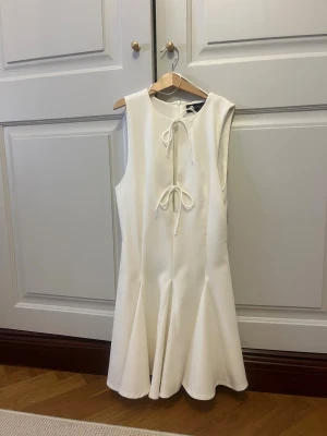Vit omlottklänning från Zara XS - Supersnygg vit kortklänning från Zara i storlek XS. Klänningen har omlottdetaljer med två knyt framtill, är ärmlös och har en lätt utsvängd kjol. Materialet är mjukt och har en lätt struktur. Aldrig andvänt pågrund av storlek . Dragkedja i ryggen för enkel påklädning.