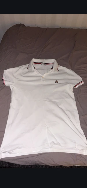 Vit Moncler pikétröja med röd detalj - Vit pikétröja från Moncler med klassisk krage och knappar framtill. Röd och marinblå rand på krage och ärmslut ger en snygg kontrast. Tröjan har Moncler-logga broderad på bröstet och är gjord i mjuk bomull som andas.