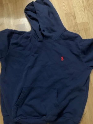 Mörkblå hoodie från Polo Ralph Lauren - Mörkblå hoodie från Polo Ralph Lauren med klassisk röd logga broderad på bröstet. Hoodien har huva, magficka och ribbade muddar vid ärmslut och nederkant. Tillverkad i mjukt bomullsmaterial som är skönt att bära.