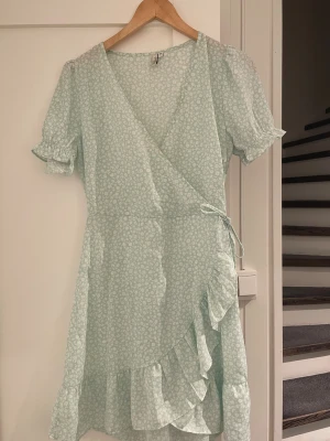 Ljusblå blommig omlottklänning NLY Trend - Supersöt ljusblå kortklänning från NLY Trend med vitt blommigt mönster. Omlottmodell med volangkant, puffiga kortärmar med rynk och knyt i midjan. Klänningen har en diskret dragkedja i sidan och är gjord i mjuk polyester. Perfekt för dig som gillar romantisk och fräsch stil.