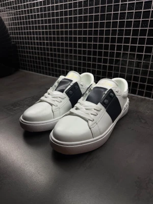 Valentino opens  - Snygga vita sneakers från Valentino Garavani med bred mörkblå läderdetalj över sidan och tungan. Skorna har rund tå, platt sula och vita snören. Märkeslogga i guld på plösen. Perfekt för dig som gillar stilrena och lyxiga sneakers.