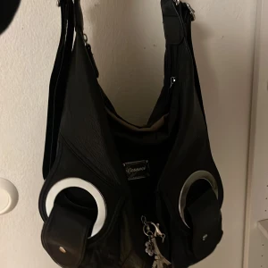 blå axelväska från Gessaci med detaljer - Cool mörkblå axelväska från Gessaci i skinn med stora silvriga ringdetaljer framtill och två fickor med lock. Väskan har en bred axelrem och dragkedja upptill. Perfekt för dig som vill ha en unik och rymlig väska med edgy vibe.