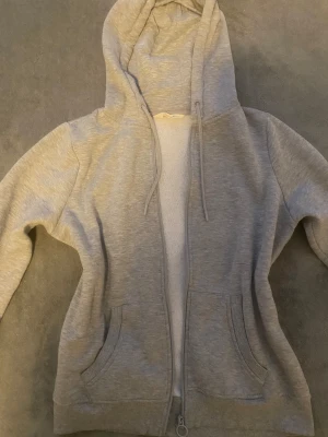 Grå hoodie med dragkedja och huva - Snygg grå hoodie med dragkedja och huva, perfekt för chill dagar. Tröjan har långa ärmar, två fickor fram och ribbade muddar vid ärmslut och nederkant. Materialet är mjukt och känns skönt mot huden. Klassisk look med snörning i huvan och rund dragkedja.