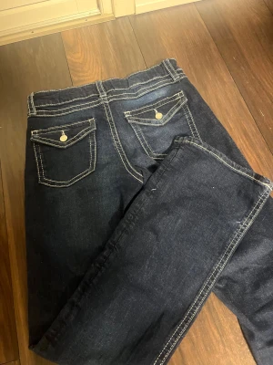 Mörkblå jeans med kontrastsömmar - Snygga mörkblå jeans med vita kontrastsömmar och fickor med lock och knapp bak. Klassisk femficksmodell med bälteshällor och dragkedja fram. Perfekta för dig som gillar en stilren och tidlös look. Den har dock lite slitna delar på slutet av jeansen på baksidan 