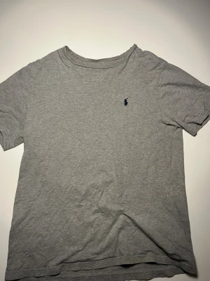 Grå t-shirt från Polo Ralph Lauren - Snygg grå t-shirt från Polo Ralph Lauren med klassisk rund hals och korta ärmar. T-shirten är i mjuk bomull och har den ikoniska mörkblå polospelaren broderad på bröstet. Perfekt basic-plagg med clean look.