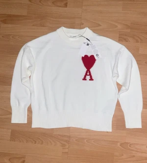 Vit ami tröja  - Vit stickad tröja från Ami Alexandre Mattiussi med röd hjärtdetalj och bokstaven A på bröstet. Rund hals, ribbade muddar och nederkant. Mjuk och skön bomullskvalitet, perfekt för dig som gillar clean stil med en twist.