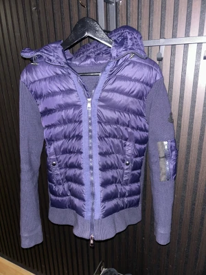 Blå cardigan  från Moncler black label - Säljer en snygg blå cardigan från Moncler med stickade ärmar och quiltad front. Den är även black label.Jackan har huva, två fickor med knappar och en extra ficka med dragkedja på ärmen. Perfekt för dig som vill ha en cool och unik jacka med både stil och funktion.