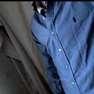 Blå skjorta från Ralph Lauren - Säljer en klassisk blå skjorta från Ralph Lauren i slim fit-modell. Skjortan har knappar framtill, lång ärm och den ikoniska broderade loggan på bröstet. Tillverkad i bomull för en skön känsla och snygg passform.