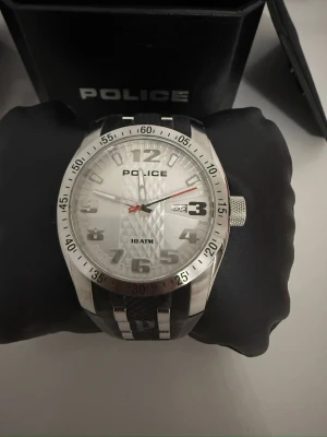 Police herrklocka i silver och svart - Snygg och robust herrklocka från Police med rund silverfärgad boett och svart armband i syntetmaterial. Urtavlan är silver med markerade siffror, datumvisning och röd sekundvisare. Vattentät upp till 10 ATM. Levereras i originalbox.